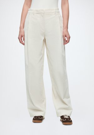 Personne portant un pantalon en velours côtelé écru à jambes larges et des chaussures décontractées marron, debout devant un fond blanc uni.