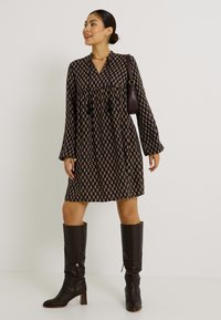 Robe noire à motifs avec des manches longues, décolleté en V et taille froncée ; associée à des bottes en cuir marron jusqu'aux genoux.