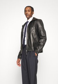 Veste en cuir noir avec fermeture �éclair à l'avant, poignets côtelés et un design structuré, portée sur une chemise blanche et un pantalon bleu marine.