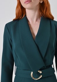 Teal wikkeljurk van gladde stof met een diepe V-hals en brede revers. Heeft een gouden ronde gesp op een bijpassende riem.