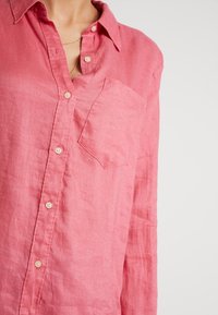 Chemise en lin rose à boutons avec col, poche poitrine unique et texture subtile. Présente des boutons de couleur claire et des manches longues.