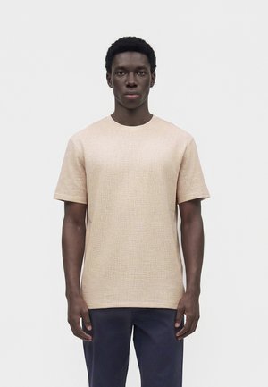 Jonge man met kort krullend haar, gekleed in een beige textuur kortemouwen overhemd en donkerblauwe broek, staand tegen een effen witte achtergrond.