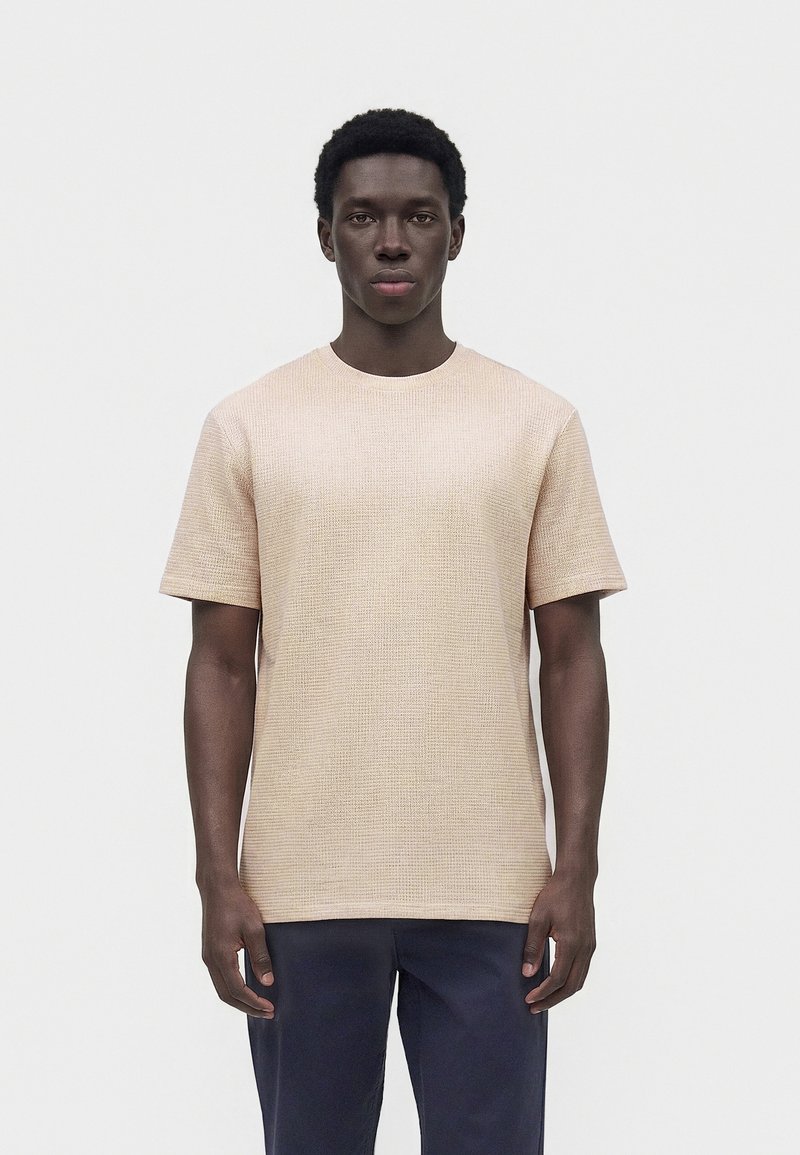 Νέος άνδρας με κοντά σγουρά μαλλιά που φοράει μπεζ υφασμάτινο κοντομάνικο πουκάμισο και σκούρο μπλε παντελόνι, στέκεται μπροστά από ένα απλό λευκό φόντο.