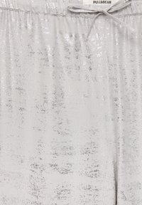 Jogging gris clair avec un léger brillant métallique, comportant une taille élastique, un cordon de serrage et une surface texturée avec des motifs éparpillés.