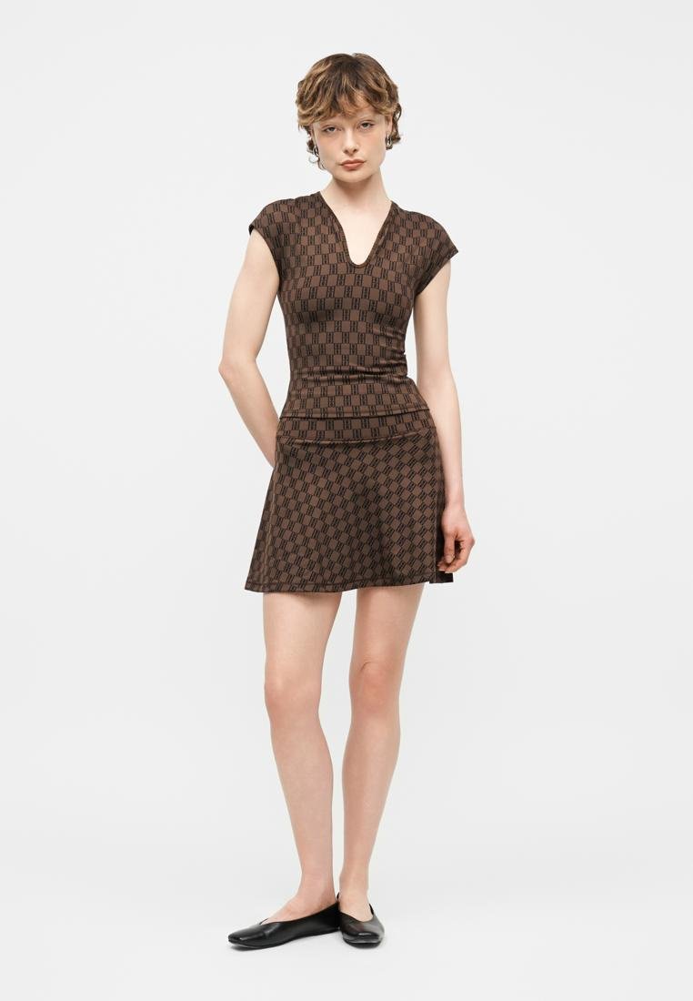 Femme debout, les mains derrière le dos, portant un haut moulant à manches courtes marron à motifs et une jupe évasée assortie, avec des ballerines noires.