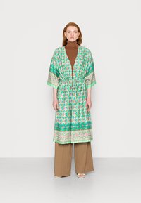 Vestido estilo kimono verde com padrão, com um laço na cintura, mangas curtas e um acabamento floral contrastante. Usado sobre um suéter de gola alta castanho e calças bejas.