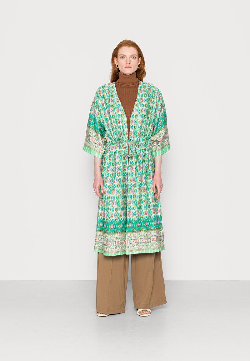 Vestido estilo kimono verde com padrão, com um laço na cintura, mangas curtas e um acabamento floral contrastante. Usado sobre um suéter de gola alta castanho e calças bejas.