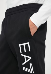 Sorte joggebukser med ribbestrikket liv, med hvit "EA" tekst og linjer på siden, samt en sidelomme. Stoffet ser glatt og mykt ut.