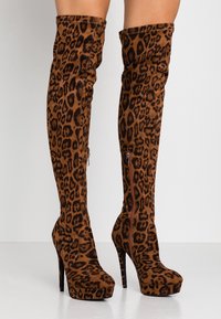 Botas de tacón alto sobre la rodilla con estampado de leopardo marrón y negro, cremalleras laterales y suelas de plataforma sobre un fondo blanco.