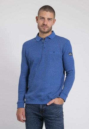 Homme aux cheveux courts et barbe portant un polo bleu à manches longues et un jean, une main dans la poche, sur un fond clair uni.