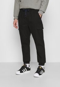 Černé pracovní kalhoty s elastickým pasem, manžetami u kotníků, bočními kapsami a šitými detaily, doplněné černo-bílými teniskami.