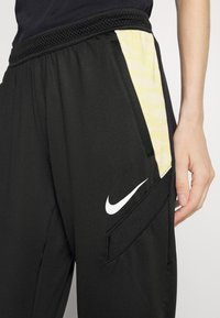 Schwarze Sporthosen mit gelbem gemustertem Seitenpanel, elastischem Bund und weißem Nike-Logo am Unterschenkel. Glatte Textur, lockere Passform.