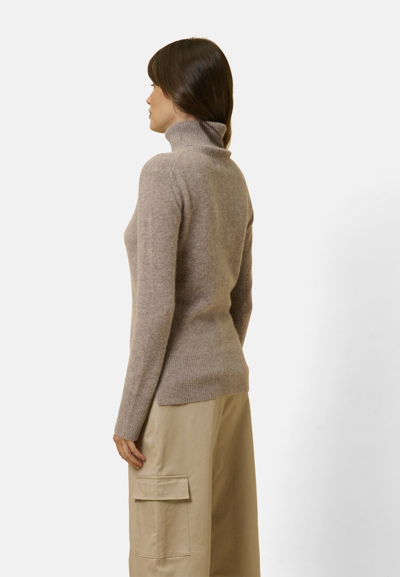 Maglione beige con collo a dolcevita in morbido tessuto, silhouette aderente, polsini e orlo a coste, mostrato di lato, abbinato a pantaloni cargo di colore chiaro.