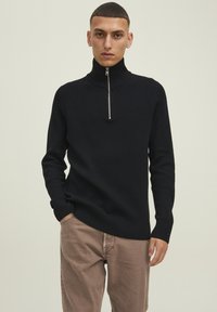 Maglione nero a costine con colletto a mezza zip, dotato di maniche lunghe e vestibilità rilassata. Indossato sopra pantaloni beige chiaro.
