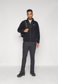 Black fleece jacket with zip, gray shirt underneath, black pants, and black hiking boots. Backpack with patterned straps.