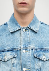 Κοντινό πλάνο ενός ατόμου που φοράει μια ανοιχτού μπλε τζιν ζακέτα με κουμπωτές τσέπες στο στήθος και μεταλλικά κουμπιά.