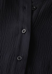 Camicetta blu scuro con texture caratterizzata da un tessuto stropicciato, con bottoni neri lungo il davanti, creando un aspetto sottile e stratificato.