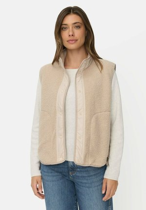 Vrouw draagt een beige mouwloos fleecevest over een lichtgekleurde shirt met lange mouwen en een blauwe spijkerbroek, staand tegen een effen witte achtergrond.