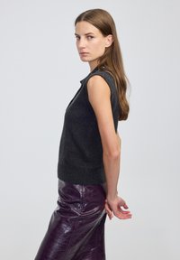 Kobieta z długimi brązowymi włosami, ubrana w bezrękawnik w ciemnoszarym kolorze i błyszcząca purpurową skórzaną spódnicę, stoi bokiem z rękami za plecami.