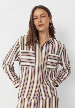 Kvinde med langt brunt hår iført guldringe i ørerne samt en brun, hvid og lilla skjorte med lodrette striber og tilhørende bukser.