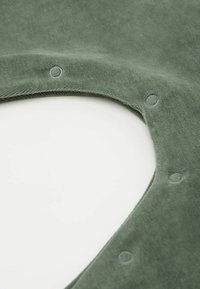 Grüner Stoff mit einer weichen Textur, der eine abgerundete Aussparung und kleine kreisförmige Akzente aufweist. Die Oberfläche ist glatt und einheitlich.