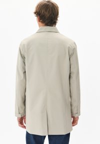 Svetlo siva dolga trench jakna z zadnjim razporkom, gumbi na manšeti in klasičnim ovratnikom, izdelana iz gladkega, vremensko odpornega blaga.