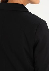 Černý blazer s vypasovaným střihem, hladkou texturou a klopovým límcem. Zobrazuje zadní část s viditelným švem.