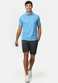 Plavi pamučni polo majica s kragnom i malim crvenim logom, u kombinaciji s sivo kratkim hlačama i bijelim tenisicama, noseći ih muški model koji stoji.
