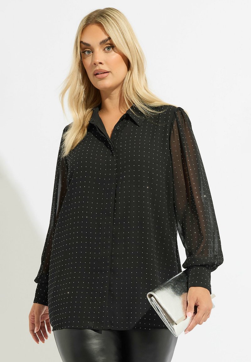 Blusa nera con maniche trasparenti, motivo a pois, chiusura frontale con bottoni e colletto a punta. Clutch argento tenuta in mano.