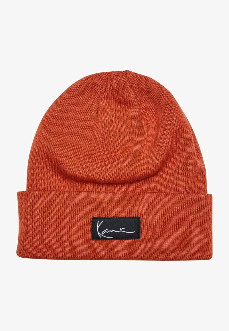 Knitted beanie in vibrant rust color, featuring a cuffed design and a rectangular black label with white script logo. Soft texture.