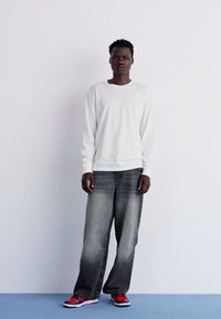 Camicia bianca a maniche lunghe abbinata a jeans grigi larghi e sneakers rosse. Design semplice e casual con tonalità neutre e accenti di calzature audaci.