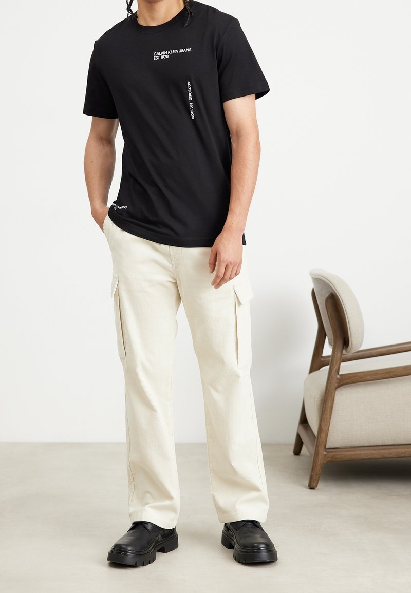 Svart bomullströja med vit text "Calvin Klein Jeans", kombinerad med krämfärgade cargobyxor med sidofickor och svarta kraftiga skor.