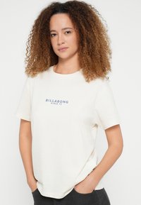 Μπεζ βαμβακερό t-shirt με κοντά μανίκια και στρογγυλή λαιμόκοψη. Εκτύπωση μπροστά: "BILLABONG SINCE 73" σε μπλε χρώμα. Απαλή υφή, άνετη εφαρμογή.