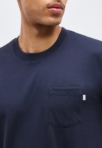 Κοντινό πλάνο ενός ατόμου που φοράει μπλε σκούρο t-shirt με στρογγυλή λαιμόκοψη, μικρή τσέπη στο στήθος και λευκή ετικέτα υφάσματος.