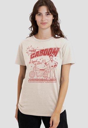 Young woman wearing beige T-shirt featuring red graphic of a cartoon man giving thumbs-up next to a motorcycle with "Duke Caboom" text.