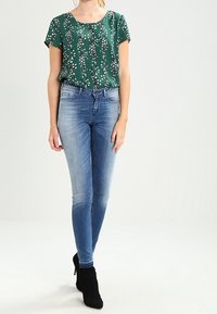 Blusa verde con estampado floral y mangas cortas, combinada con jeans ajustados de color azul claro y botines negros. Tejido suave y corte casual.