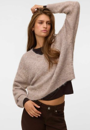 Jeune femme aux longs cheveux bruns portant un pull beige duveteux par-dessus un haut noir et un pantalon foncé, posant avec un bras levé derrière la tête.