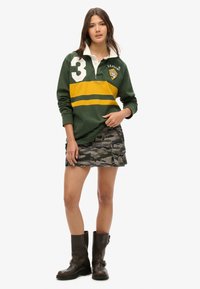 Grünes Rugby-Polo mit weißem Kragen, gelben und grünen Streifen und der Nummer 3; kombiniert mit einem Camouflage-Minirock und braunen Stiefeln.