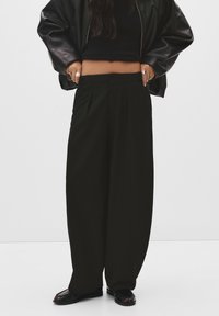 Črne široke hlače z gubami, nosene z krojenim črnim topom in velikim črnim plaščem. Gladek material, minimističen dizajn in brez vidnih vzorcev.