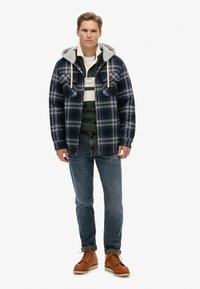 Ternede flanel skjorte i marineblå og hvid, lagd over en mørkegrøn top. Båret med blå jeans og brune snørebåndsstøvler. Har to brystlommer.