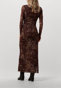 Frau in einem langärmeligen, knöchellangen, braun-beigen Leopardenmuster-Kleid mit schwarzen Knöchelstiefeln, die mit dem Rücken zu uns auf einem weißen Hintergrund steht.