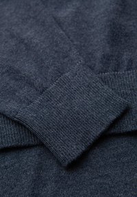 Primo piano di un maglione blu scuro, caratterizzato da un knit testurizzato con polsini a costine e una finitura morbida. Il tessuto appare leggero e flessibile.