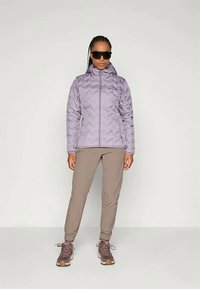 Ženska v vijoličnem podloženem jopiču, bež hlačah, rjavih pohodniških čevljih in velikih črnih sončnih očalih, stoji proti enostavni svetli ozadju.