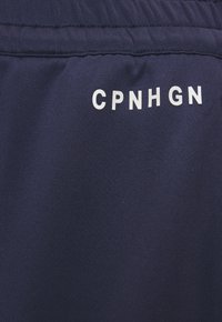 Pantalones cortos deportivos de color azul marino hechos de tela texturizada, con un logotipo impreso en blanco "CPNHGN" en la parte posterior. Incluyen una cinturilla elástica.