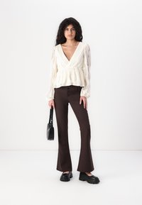 Hvit peplum-bluse med teksturert stoff og lange ermer, sammen med brune vide bukser og svarte kraftige platå-sko, holder en liten veske.