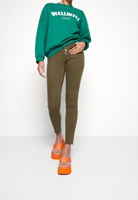 Grön sweatshirt med vit text, olivgröna slim fit jeans och orange plattformssandaler. Har tre knappar vid midjan och trasiga ankleder.