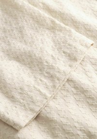 Κρεμ ύφασμα με ανάγλυφο υφαντό μοτίβο μικρών τετραγώνων και διακριτικές διαγώνιες πτυχές.