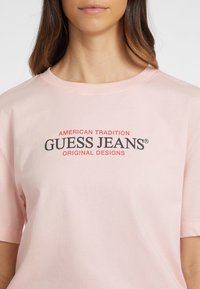 Heledane roosa puuvillane T-särk lühikeste varrukatega, millel on mustade ja punaste kirjatega tekst "GUESS JEANS" ning "AMERICAN TRADITION" punases kirjas selle üle.