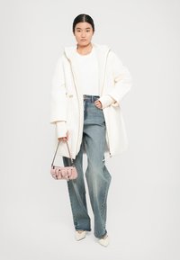 Witte oversized jas met een capuchon, gecombineerd met een witte top en lichtblauwe jeans. Een kleine roze handtas en puntige witte schoenen maken de look compleet.