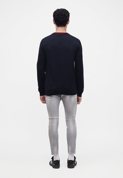 Homme debout de dos, portant une chemise noire à manches longues, un jean gris clair, des chaussettes blanches et des baskets noires sur fond uni.